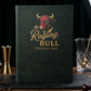 A dark green hardcover book or menu standing upright on a wooden table, featuring a red bull illustration and the text 'the Raging Bull COCKTAIL BAR' in gold. Bar-themed props including a jigger, glass, and cherry are visible around it against a dark backdrop.