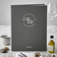 A dark gray hardcover menu book for Herefordshire High Care Homes, styled on a white marble table with silver cutlery, a small bottle of olive oil, a wine glass, and lavender sprigs.
